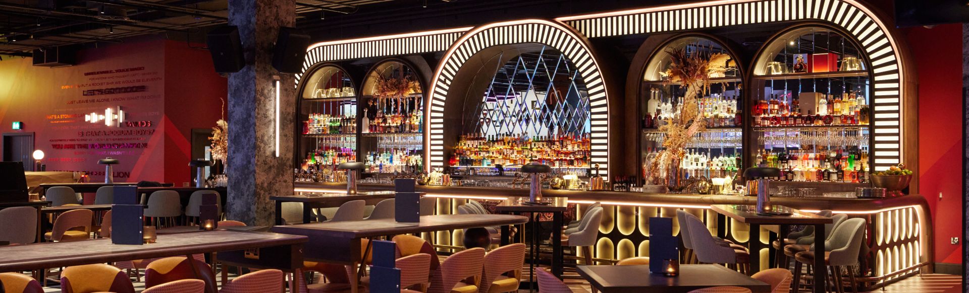Colourful bar counter, tables and chairs with red-painted walls and white neon lights above the bar.