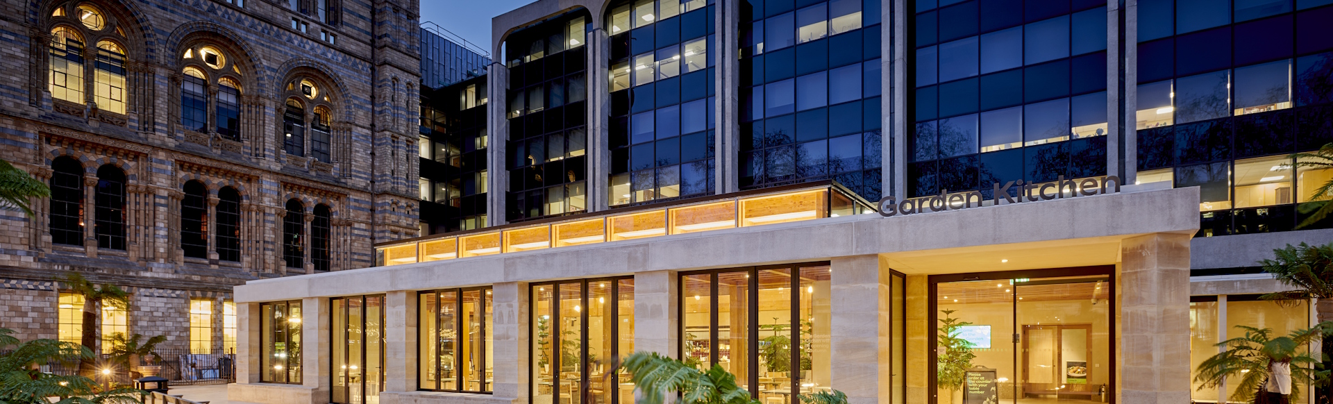 Garden Kitchen venue lit up in the evening with yellow light, with Natural History Museum building in the background to the left, modern building to the right.