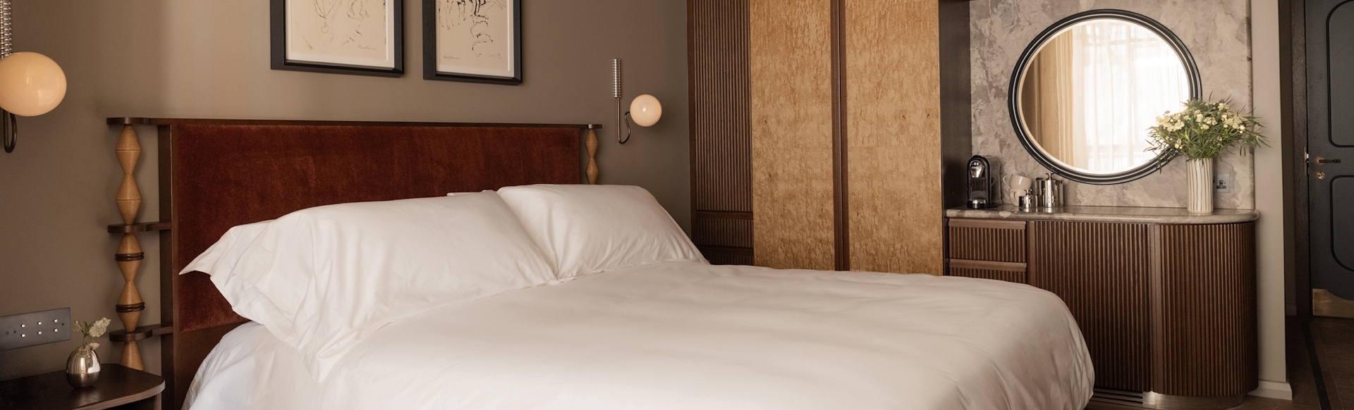 Hotel bed with white sheets, surrounding by contemporary room furnishings, a mirror and pictures on the wall.