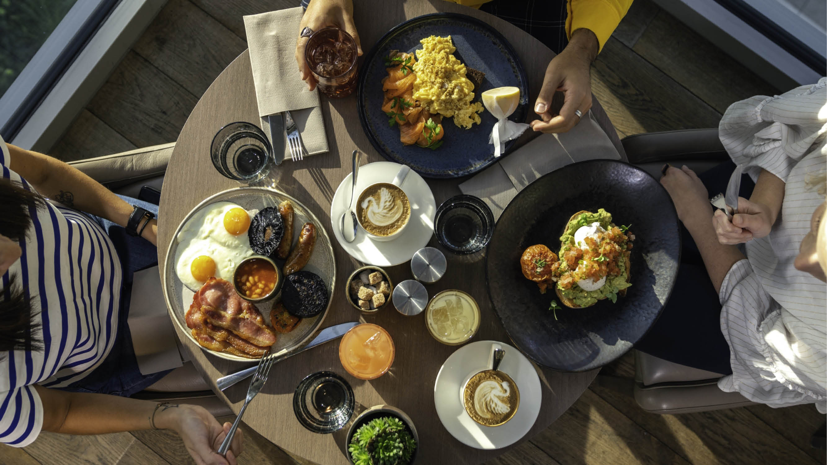 Three brunch plates, coffee and drinks placed on a wooden table, photographed from above.