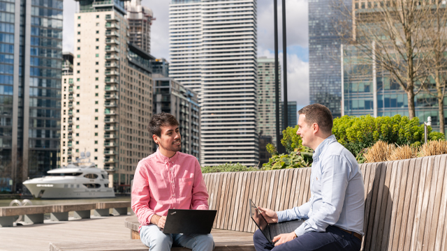 Two people with laptops sitting on a bench outside on a sunny day among office buildings and a riverboat in the background.