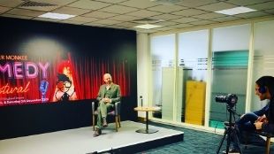 A  man in a grey suit sits in a chair on a small studio stage at CCT Venues in London.