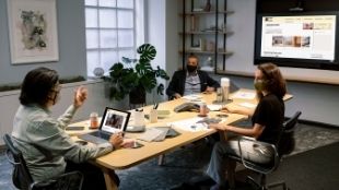 Three people sit at a wooden table with laptops and work material in London.