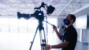 A man wearing a face mask is working behind a film camera in a studio with bright natural light.
