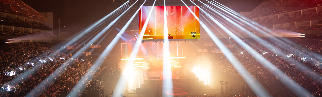 A view of an arena full of people with a screen and spotlights pointing towards the audience in the centre of the arena.