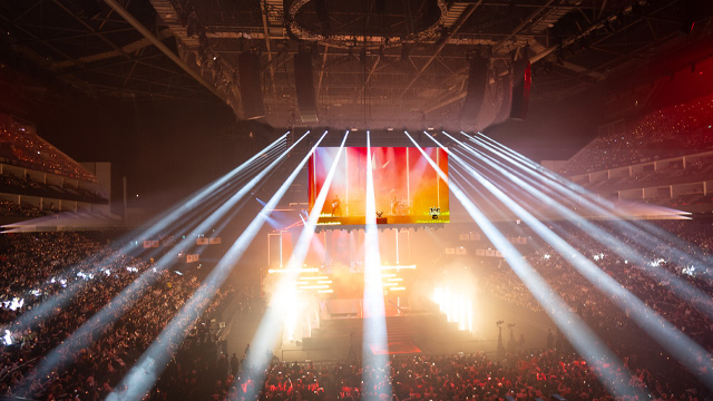 A view of an arena full of people with a screen and spotlights pointing towards the audience in the centre of the arena.