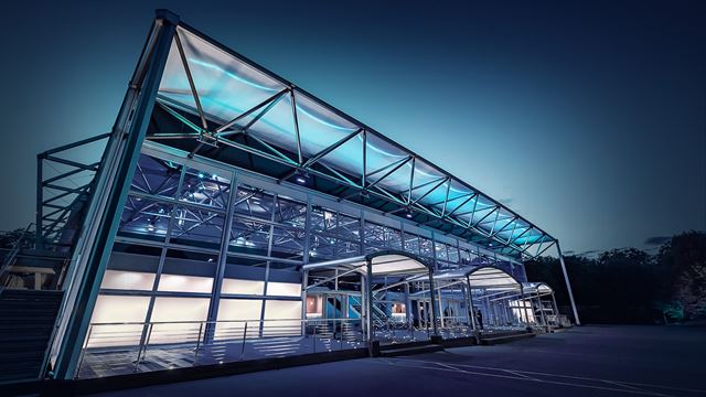 Evolution London is a versatile venue for events. Credit: Evolution London. Image courtesy of Evolution London. Steel and glass structure of Evolution London events venue at night with blue and white lights.