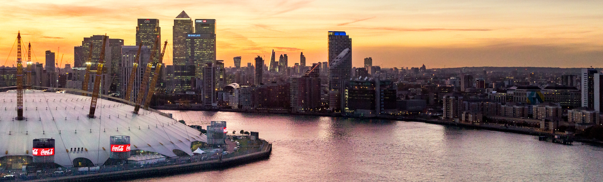 View of Canary Wharf skyline at sunset with The O2 entertainment venue and the river Thames in the foreground.