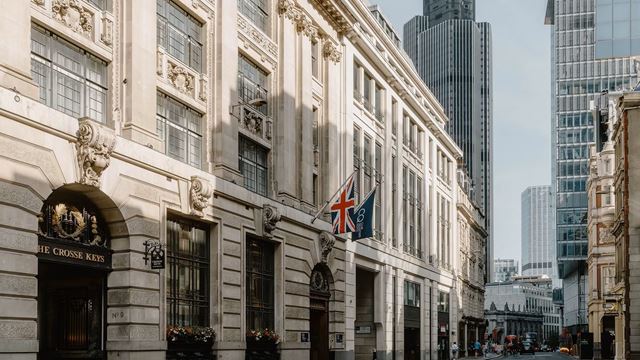 Host an event or enjoy a stay at Club Quarters London City. Credit: Club Quarters. Image courtesy of Club Quarters. Exterior of Club Quarters hotel in the City of London with the UK and hotel flags above the entrance and skyscrapers and blue sky in the background.