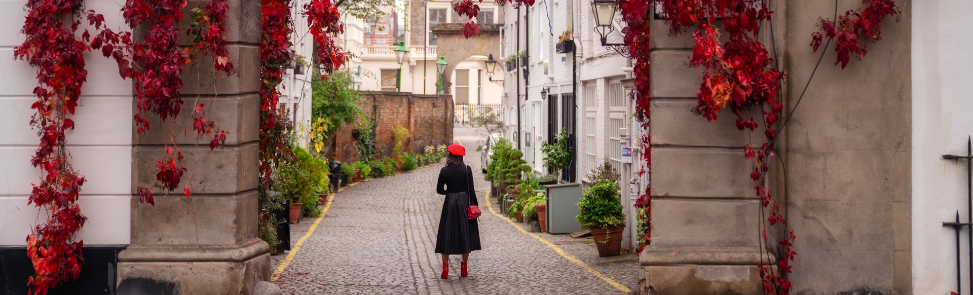 A woman dressed in a black coat and a red hat standing in front of Kynance Mews in Kensington, with red flowers hanging from the archway.