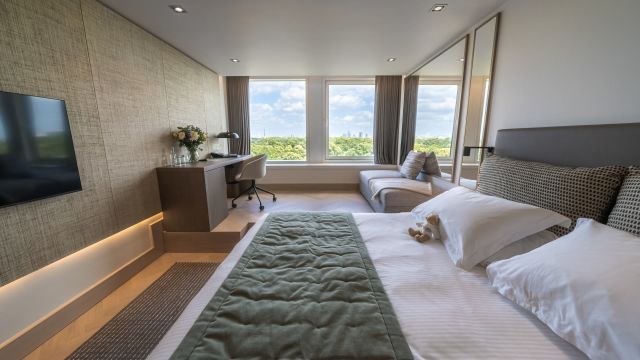 Large hotel room with white and green sheets, wooden floors, a TV and lounge area with big windows overlooking Hyde Park.