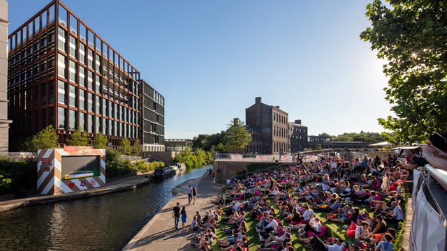 People sitting in the sun beside a canal with people walking on a towpath.