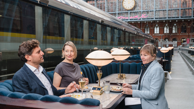 Three people in business attire sitting at a table talking and eating inside a rail station.