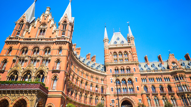 A red-brick, neo-gothic building with two towers, multiple spires and a bright blue sky in the background.