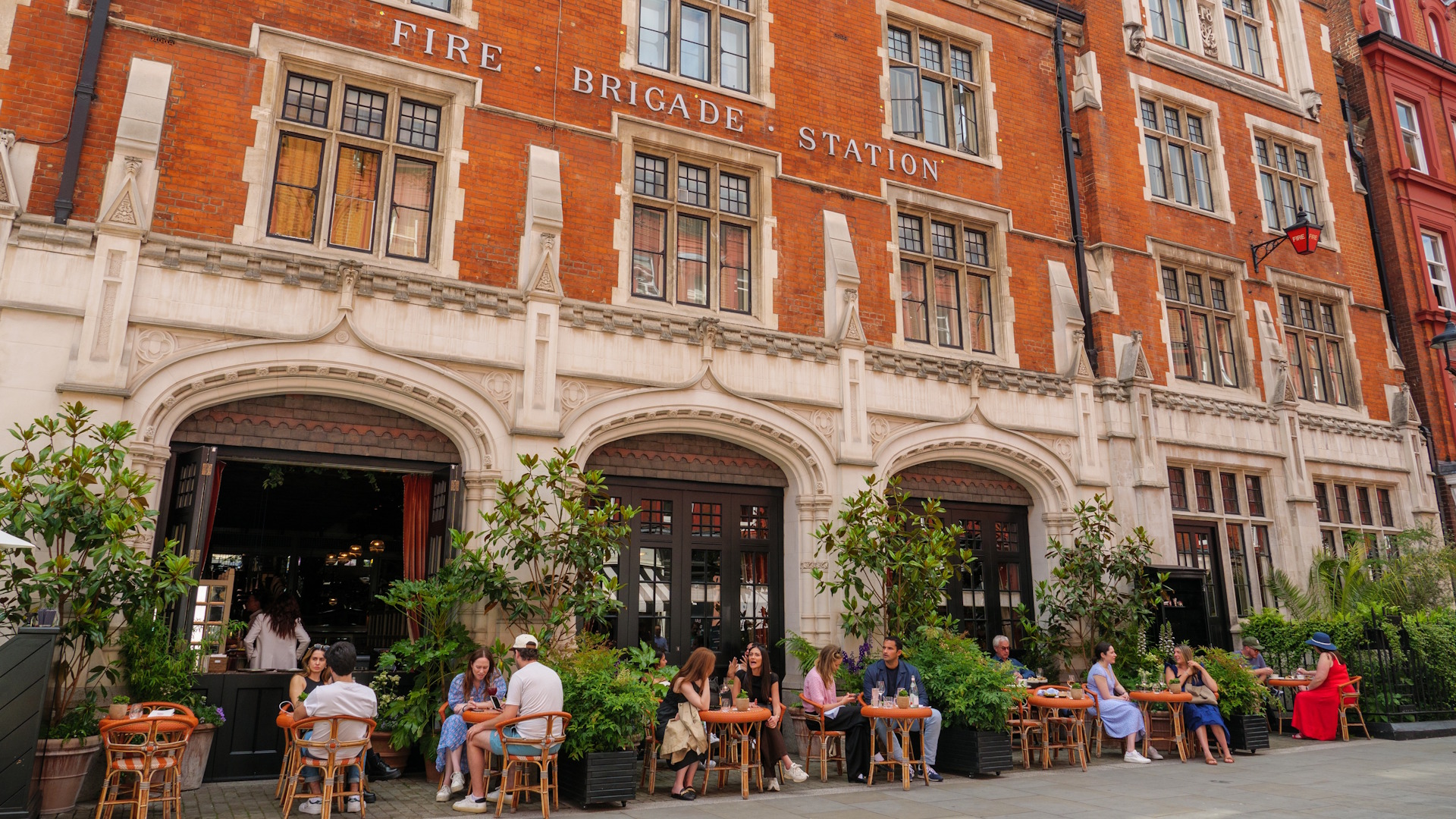 People sitting at outside tables outside red-brick restaurant building with Fire Brigade Station written on it on a summer's day.