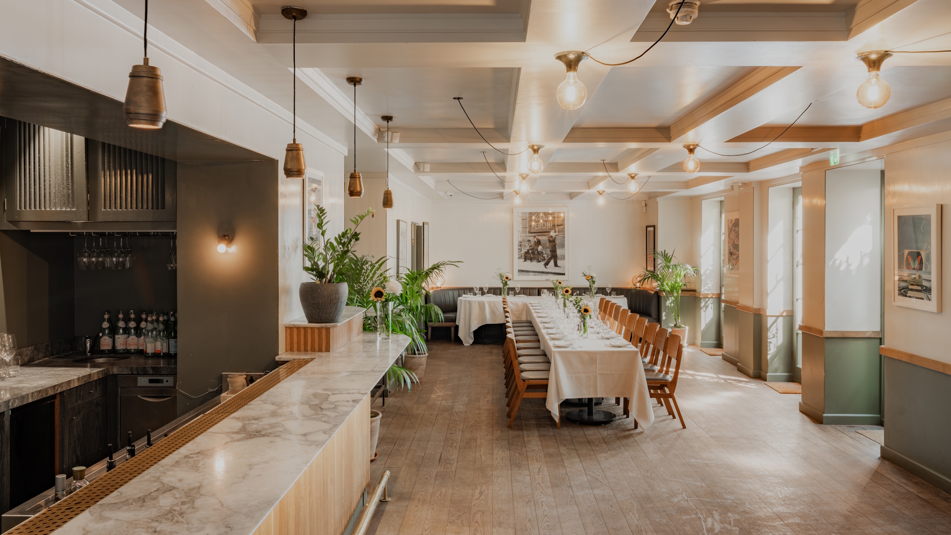 Dining room with open kitchen counter, long tables and wooden chairs, bright with natural light.