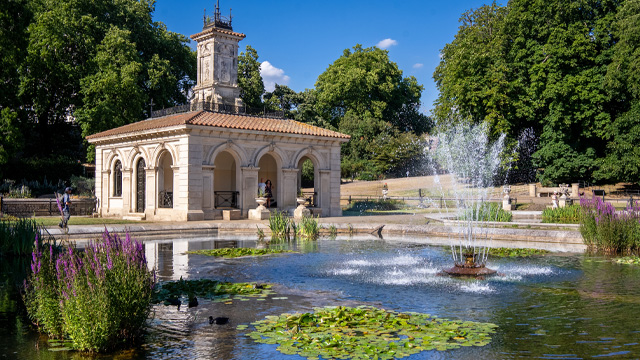 A sunny daytime view of a pond in a park with a fountain in the middle, waterlilies and a historic building surrounded by trees in the background.