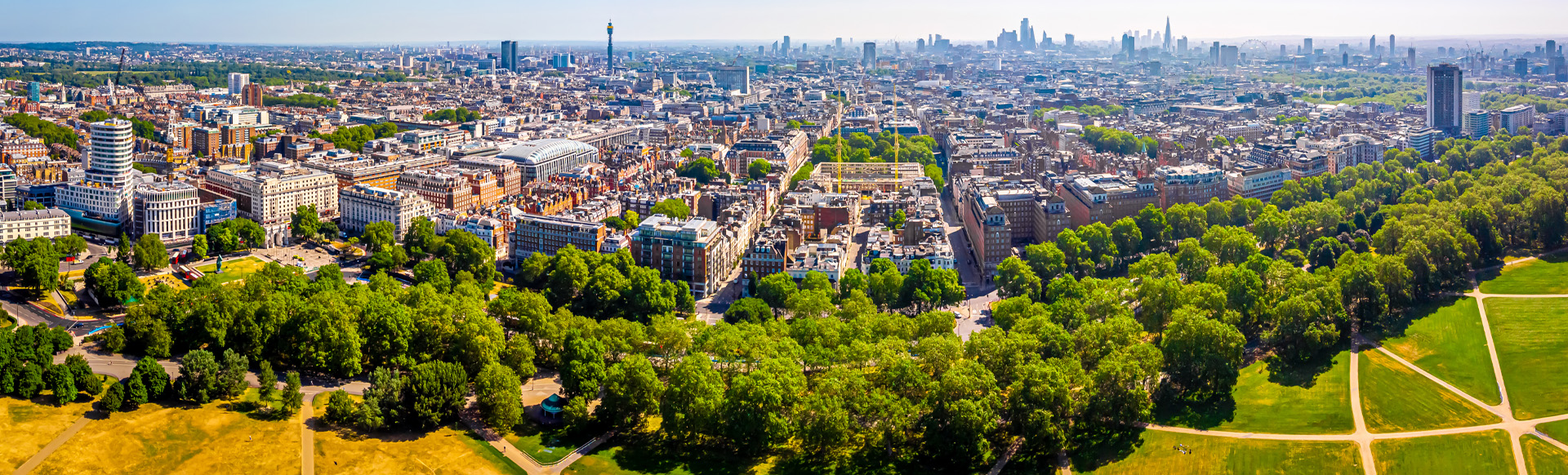 A wide angle aerial view of a central London neighbourhood with a large park at the foreground and the city skyline in the background. 