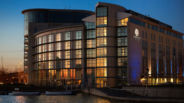 An evening view of a modern London hotel on the riverside with two boats docked in the foreground.