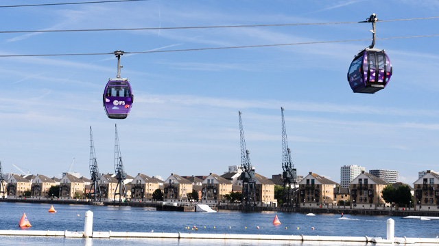 A daytime view of two purple cable cars crossing a river with houses and cranes in the background.