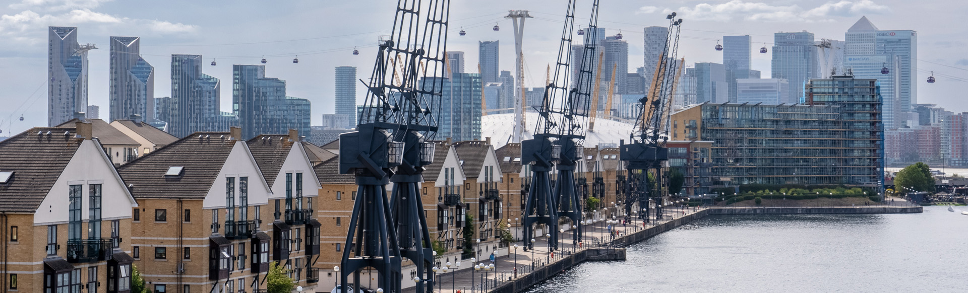 A daytime view of a row of houses on the riverside with industrial cranes on the water's edge and the skyscrapers of Canary Wharf in the background.