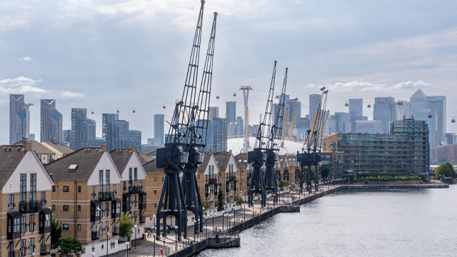 A daytime view of houses on the riverside with industrial cranes at the water's edge and the skyscrapers of Canary Wharf in the background.