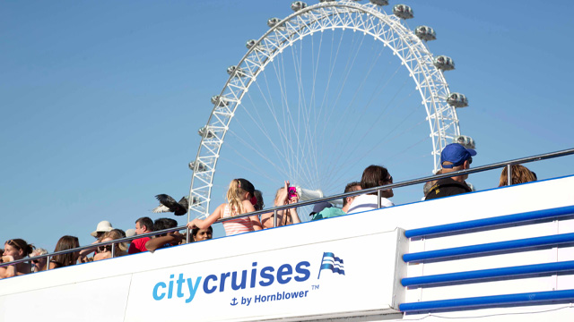 A boat with people on an open rooftop seating with the London Eye in the background.