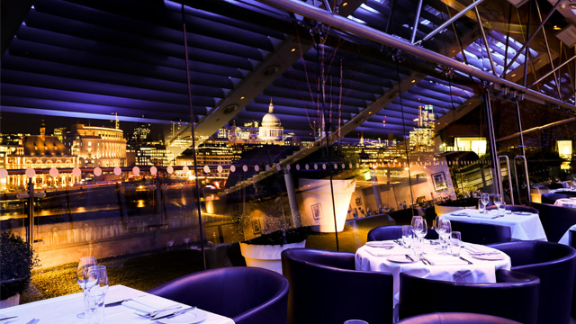 An elegant restaurant with floor to ceiling windows overlooking London at night.