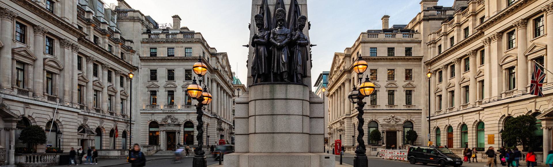 A daytime view of a London street with historic buildings and a military monument in the foreground.