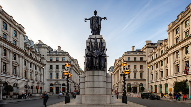 A daytime view of a London street with historic buildings and a military monument in the foreground.