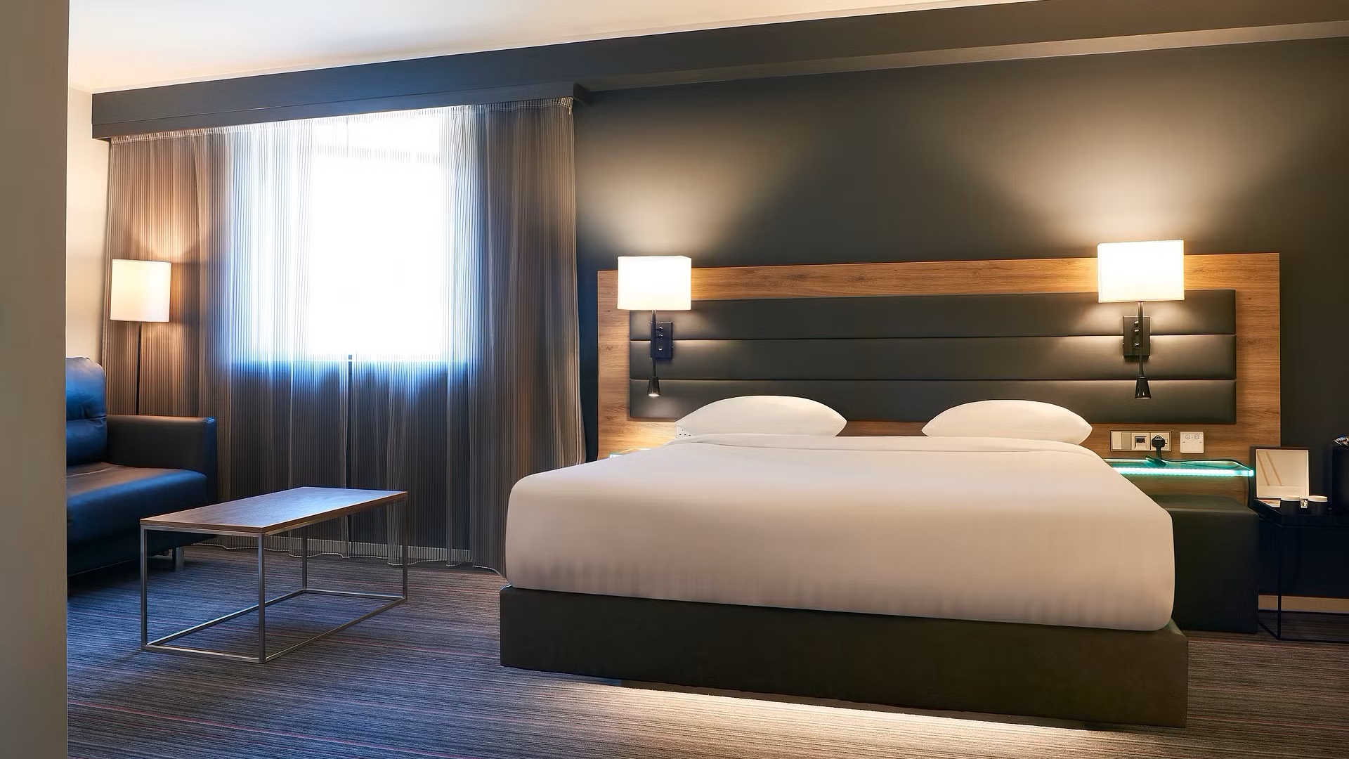 Double bed with white sheets, intimate lighting and dark walls at Moxy London Stratford.