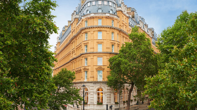 A day time view of a historic building on the corner of two streets surrounded by trees. The building has a sign that reads Corinthia London.