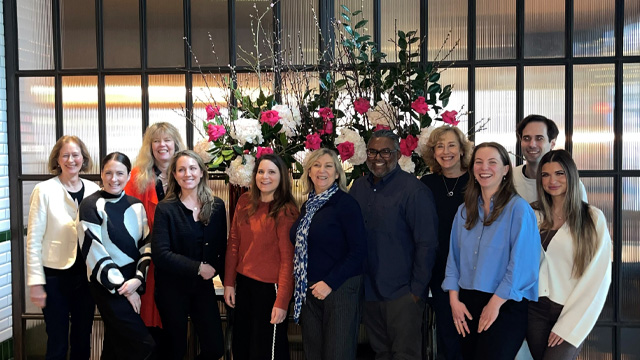A group of people in business attire posing and smiling for the camera with a tree and large window in the background.