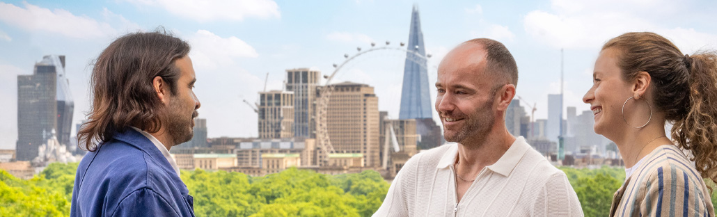 Three people smiling and talking on a rooftop terrace with views of the London skyline in the background.