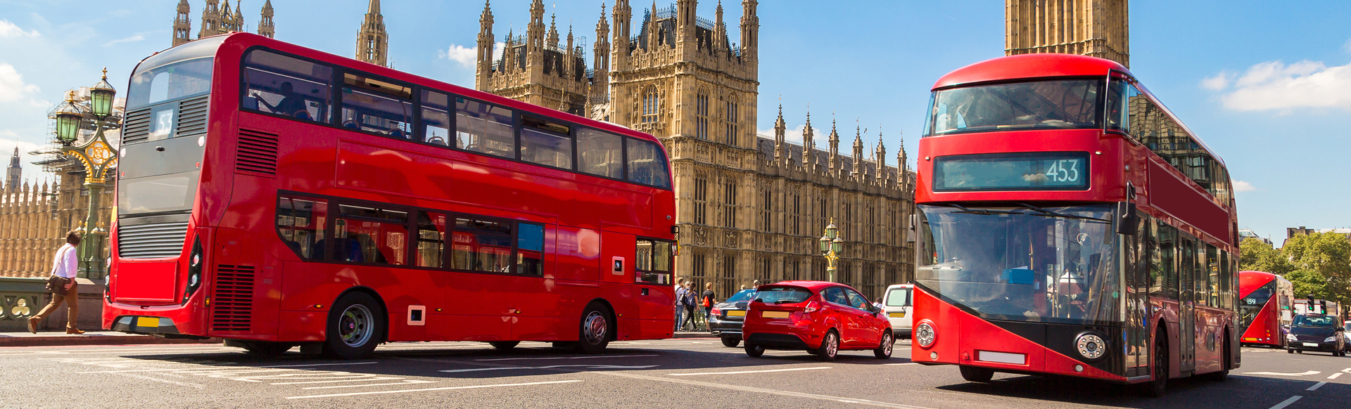 Two red double-decker buses drive in front of the Houses of Parliament in London on a sunny day.
