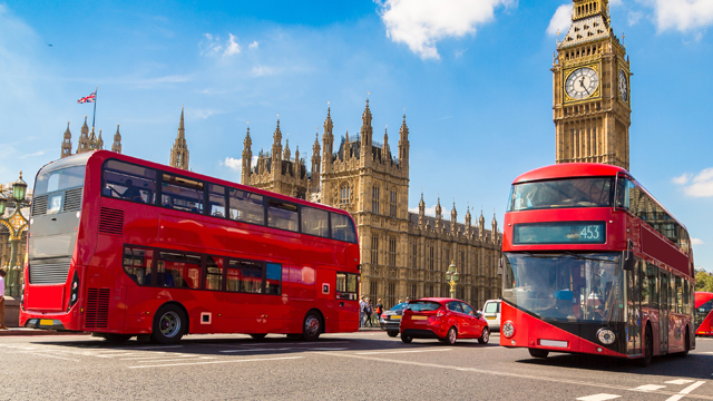 Two red double-decker buses drive in front of the Houses of Parliament in London on a sunny day.