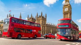 Two red double-decker buses drive in front of the Houses of Parliament in London on a sunny day.