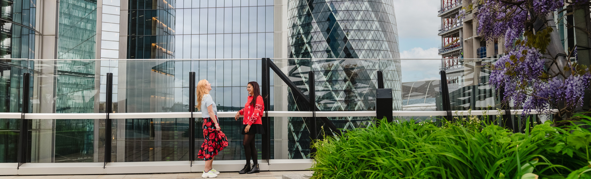 Two women talking and laughing in a rooftop garden overlooking modern skyscrapers.