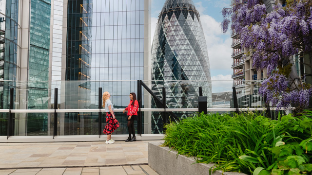 Two women talking and laughing in a rooftop garden overlooking modern skyscrapers.