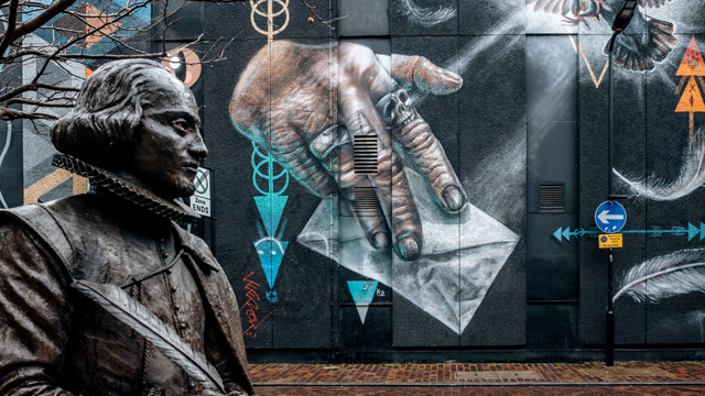 A statue of William Shakespeare with a wall covered in colourful street art in the background.