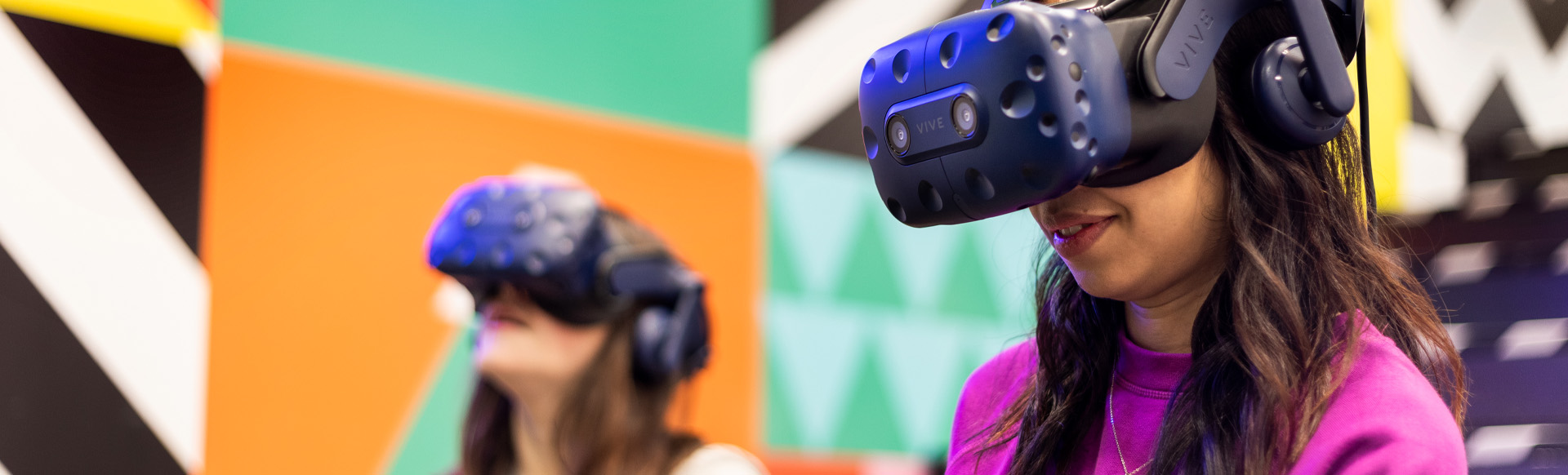 Two women wearing virtual reality headsets with colourful geometric patterns on the wall in the background.
