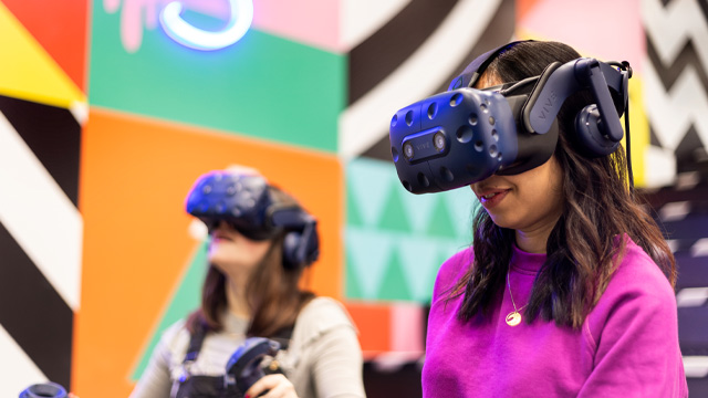 Two women wearing virtual reality headsets with colourful geometric patterns on the wall in the background.