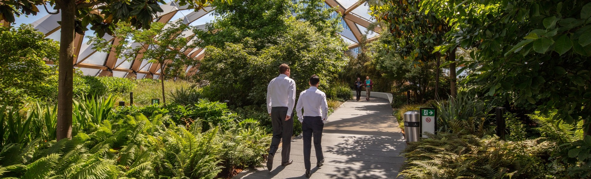 Two people walk along a path lined by green plants at the Crossrail Place Roof Garden in London.