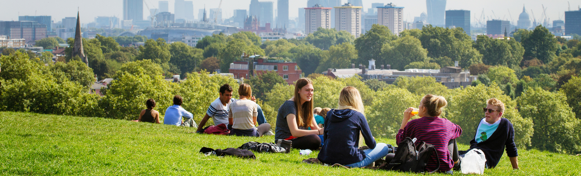 People sitting at Hampstead Heath with the London skyline in the background on a sunny day.