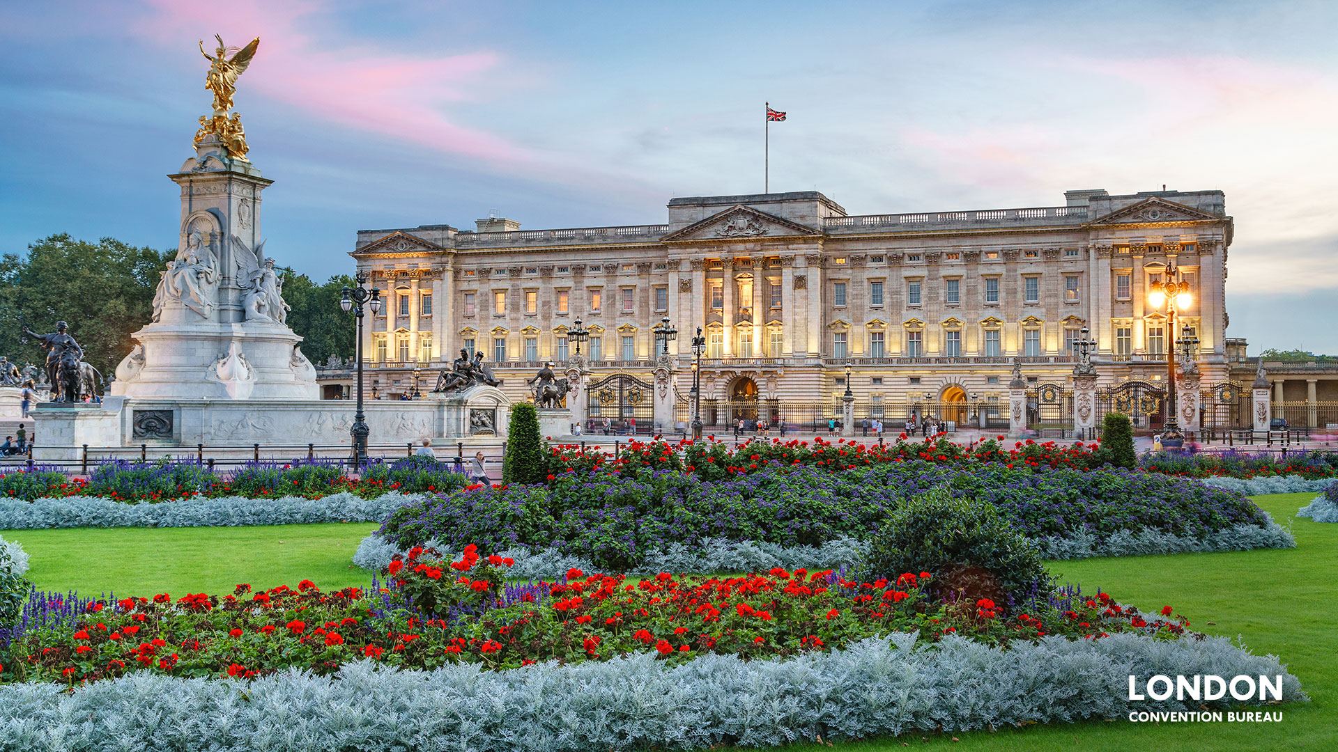 Buckingham Palace lit up behind the green garden area with red and purple flowers blooming.